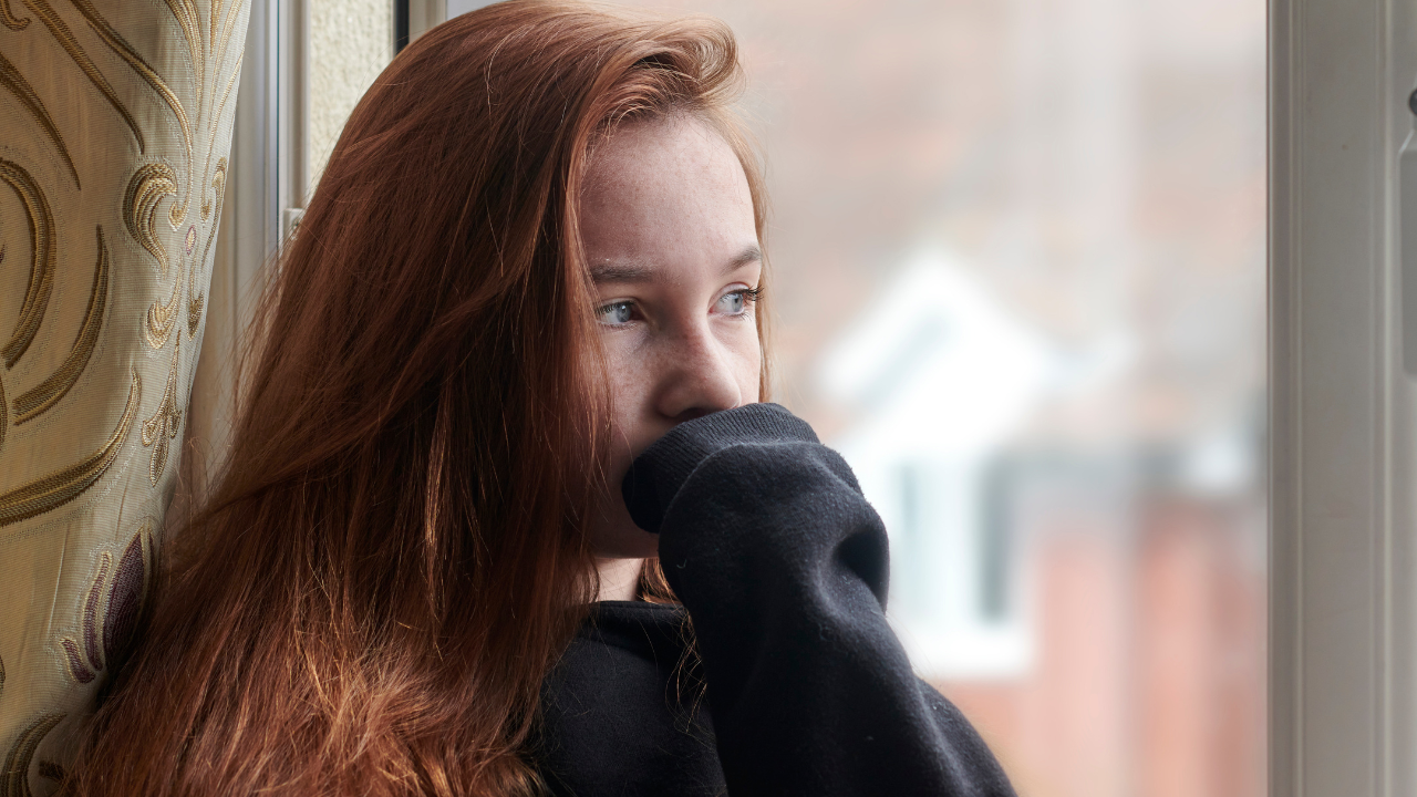 Young teen girl brooding and looking out of a window, illustrating the emotions involved when a teenager refuses to visit a non-custodial parent.